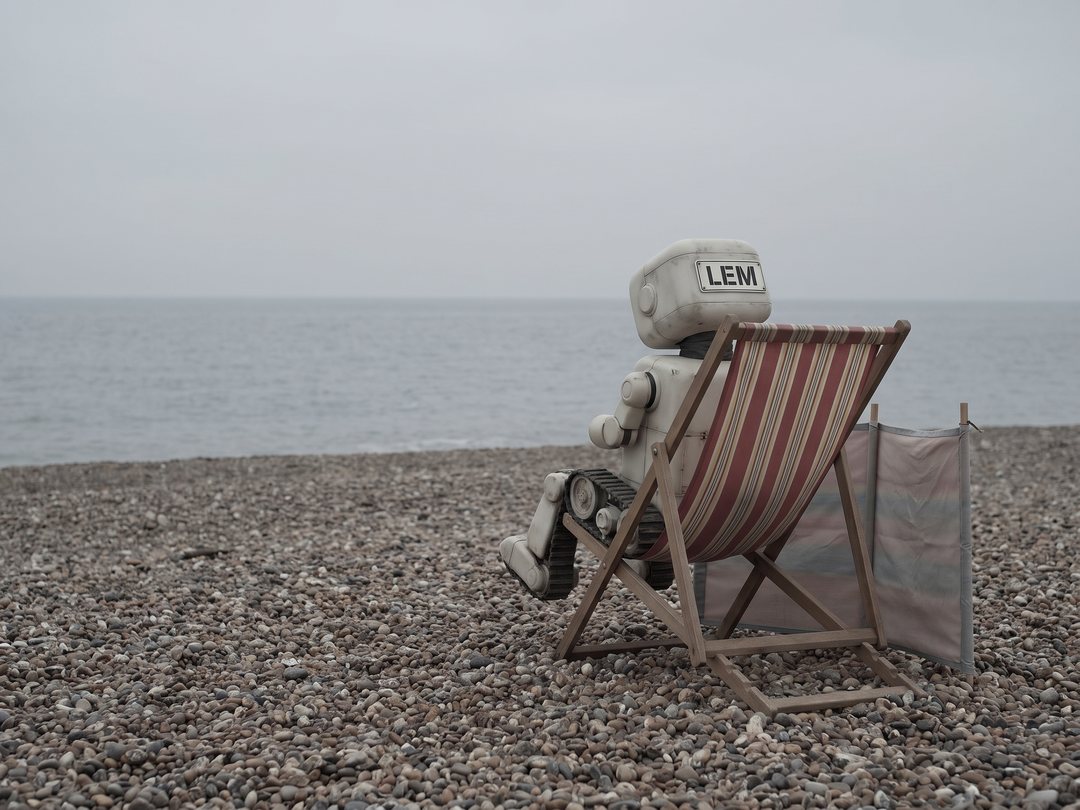 Lem, a small boxy robot with a large LEM name plate across their back, sits beside a striped deckchair on a grey pebble beach under a flat overcast sky, facing a calm sea, with a windbreak planted in the stones.