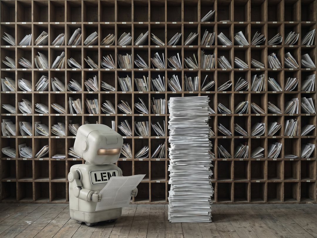 Lem, a small boxy robot with a large LEM name plate across their front, reads a single opened letter in front of a floor-to-ceiling wall of pigeonholes stuffed with post, beside a stack of unopened letters taller than they are.